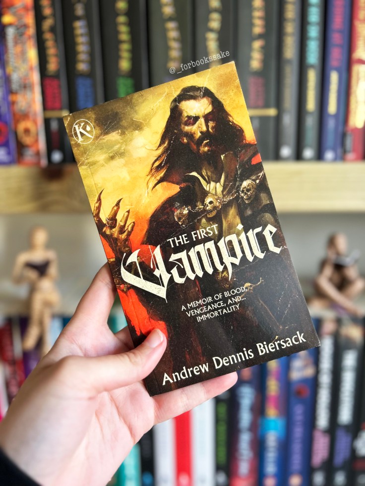 The First Vampire by Andrew Dennis Biersack paperback, held in front of blurred book shelves.
